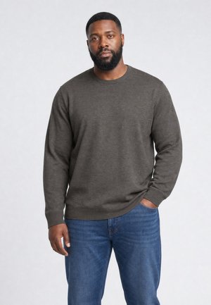 Mann mit Bart, der einen dunkelgrauen Pullover und blaue Jeans trägt, steht mit einer Hand in der Tasche vor einfarbigem Hintergrund.