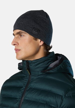 Dark gray knitted beanie with a ribbed texture and a round black logo patch. Worn with a green insulated jacket featuring a high collar.