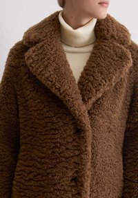 Cappotto marrone con texture, colletto a contrasto, chiusura con bottoni e tessuto peloso. Indossato sopra un maglione a collo alto color crema.
