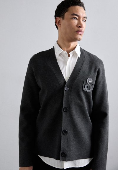 Cardigan grigio scuro con scollo a V, cinque bottoni e una patch di polpo testurizzata sul lato sinistro, indossato sopra una camicia bianca a righe.