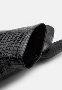 Bottine en cuir verni noir avec un motif texturé croco, présentant une ouverture arrondie et une doublure intérieure lisse.