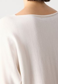 Gros plan du haut du dos et du cou d’une personne portant une chemise blanche unie avec un large décolleté, cheveux courts et bruns visibles.