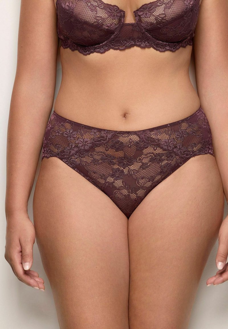 Donna con reggiseno in pizzo viola abbinato e perizoma a vita alta, fotografata dalle spalle alle cosce superiori su uno sfondo neutro.