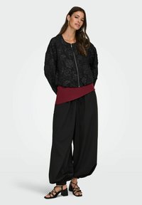 Črna teksturirana bomber jakna s cvetličnim vzorcem, preplastena preko burgundskega zgornjega dela in črnih širokih hlač, v kombinaciji s črnimi na peto vezanimi salonarji.