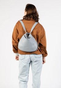 Grauer Leder-Rucksack mit runder Form, verstellbaren Trägern und Kordelzugverschluss, getragen über einer braunen Jacke und hellblauen Jeans.
