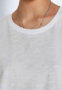 T-shirt bianco a maniche corte realizzato in tessuto leggero con una texture morbida e un colletto rilassato, indossato con più catene sottili.