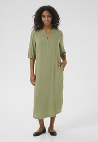 Robe longue vert olive en tissu léger. Présente un décolleté en V discret, des manches courtes et des poches latérales. Des chaussures noires plates complètent le look.