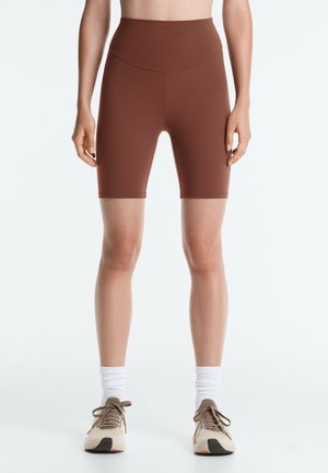 Personne portant un cuissard marron taille haute, des chaussettes blanches montantes et des chaussures de sport beiges, debout sur un fond blanc.