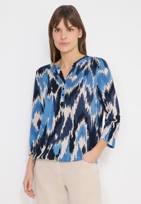 Femme portant un chemisier à motifs abstraits bleus, noirs et beiges avec des manches trois-quarts, et un pantalon beige, debout avec une main dans la poche.