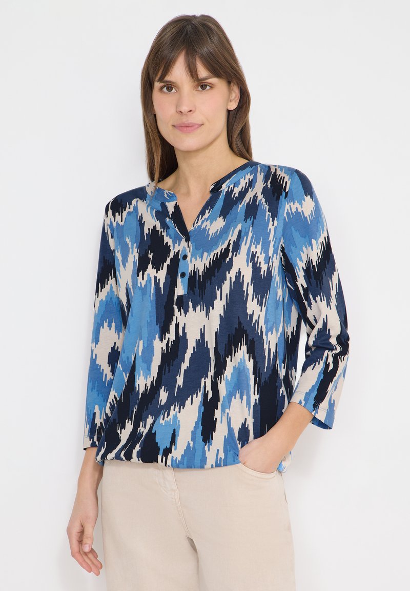 Femme portant un chemisier à motifs abstraits bleus, noirs et beiges avec des manches trois-quarts, et un pantalon beige, debout avec une main dans la poche.