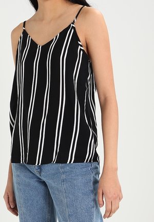 Mujer con una camiseta negra sin mangas con rayas verticales blancas y jeans de mezclilla azul, de pie contra un fondo claro y liso.