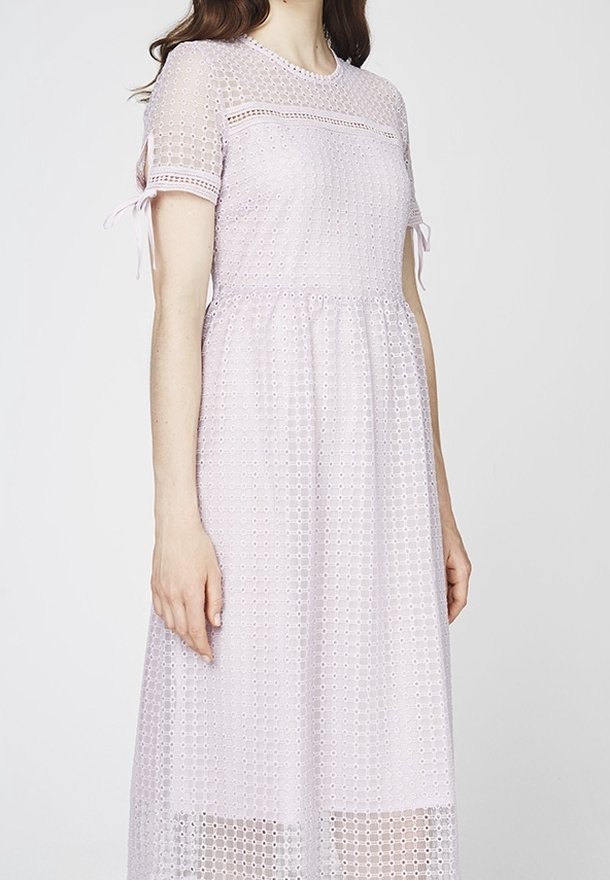 Femme portant une robe violette claire, à manches courtes et longueur genou, en tissu ajouré avec petits liens sur les manches.