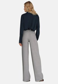 Kobieta z blond włosami, ubrana w ciemną bluzkę z długim rękawem włożoną w spodnie o wysokim stanie i szerokich nogawkach w szary kraciasty wzór, stojąca tyłem.