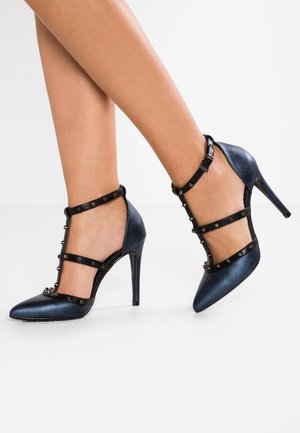 Niebieskie szpilki z ostro zakończonym noskiem, z wieloma czarnymi paskami ozdobionymi metalowymi akcentami z przodu. Gładka tekstura.