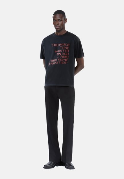 Sort bomuld t-shirt med rød tekstsætning på fronten, paired med sorte straight-leg bukser og sorte sko. Simpelt design, afslappet pasform.