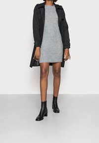 Svart trenchcoat över en grå ribbad stickad klänning, matchat med svarta ankelstövlar. Slät kvalitet, strukturerad design, åtsittande silhuett.