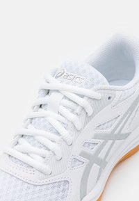 Valkoinen Asics-urheilujalkine, jossa on verkkopäällinen, valkoiset nauhat ja oranssi pohjan reuna, näkyy ylhäältä lähellä.