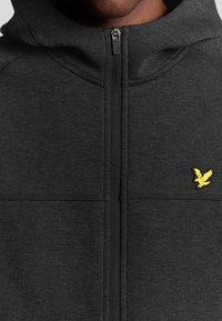Felpa con zip di colore grigio scuro, realizzata in un materiale morbido, con un piccolo logo giallo a forma di aquila sul lato sinistro del petto e dettagli delle cuciture ben visibili.