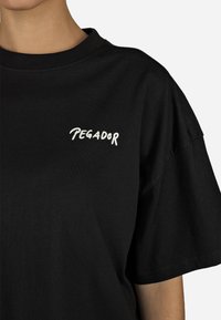 Czarna bawełniana koszulka z okrągłym dekoltem i krótkimi rękawami. Na lewej piersi widoczny napis "PEGADOR" w kolorze białym.