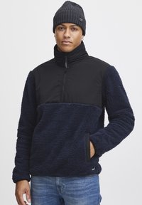 Navy fleece pullover with textured, plush fabric and black nylon panel at the neck. Features zip closure and side pockets.