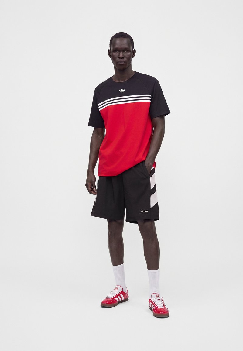 Mand iført en sort og rød Adidas-shirt, sorte shorts med hvide striber, hvide sokker og røde og hvide Adidas-sneakers, står med den ene hånd i lommen.