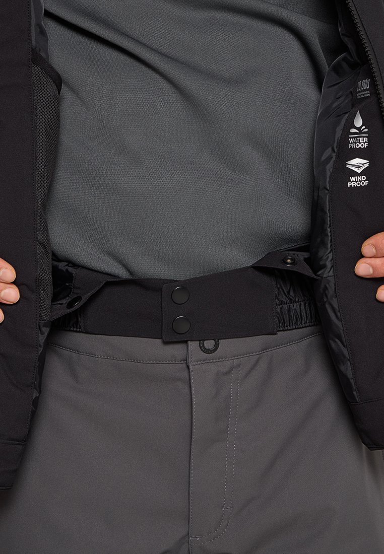 Hände, die eine schwarze Jacke mit wasser- und winddichten Etiketten anpassen, über einem grauen Hemd und grauen Hosen, wobei der Fokus auf dem inneren Bund der Jacke liegt.