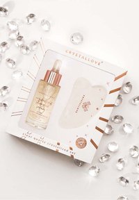 Ensemble de quartz clair cristallisé dans une boîte blanche, comprenant un flacon de sérum orné d'accents dorés et un outil lisse en forme de cœur, entouré de gemmes transparentes.