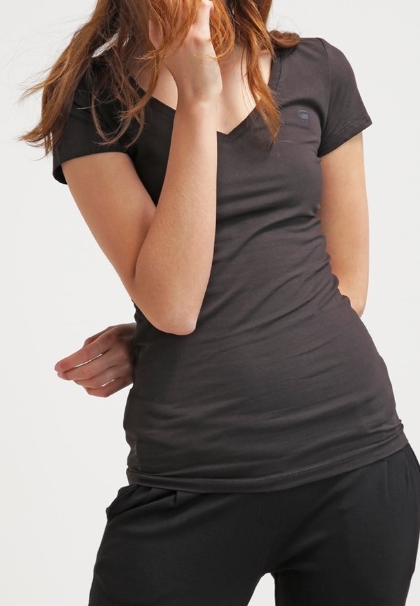 Kvinna i en åtsittande svart V-ringad T-shirt och svarta byxor, med ena handen som rör vid hennes hår och den andra bakom ryggen.