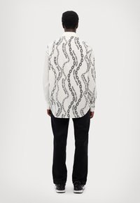 Camisa blanca de manga larga con patrón de cadenas negras, que presenta la palabra "Versace" repetidamente, combinada con pantalones negros y zapatillas.