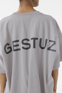 Maglietta oversized grigia con colletto rotondo. Il retro presenta un testo nero audace che dice "GESTUZ" nella parte superiore. Texture del tessuto morbida.