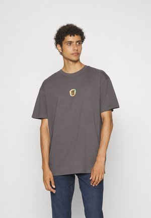 Gray cotton T-shirt with a relaxed fit, featuring a small embroidered logo in earthy tones on the chest. Short sleeves and round neckline.
