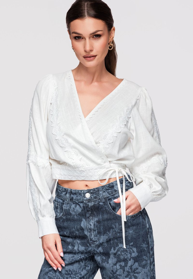 Femme portant un chemisier cache-cœur blanc orné de dentelle avec manches longues et un jean foncé à taille haute avec motif floral, une main dans la poche.