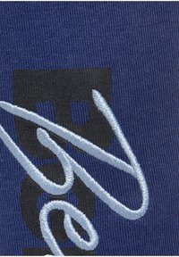 Navyblauwe stof heeft zwarte grafische tekst met geborduurde lichtblauwe accenten en draaiende lijnen, wat een textuur- en gelaagd ontwerp toont.