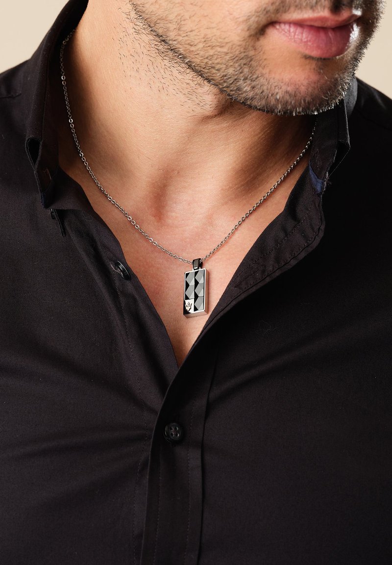 Metal halskæde med en rektangulær sort vedhæng med et geometrisk mønster og en sølvkæde, båret over en sort skjorte.