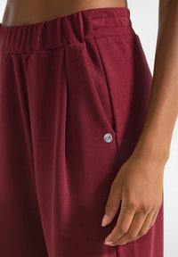Bordeauxkleurige broek van zachte stof, met geplooid ontwerp, elastische tailleband en een klein logopatch op de zijzak. Vlotte textuur.