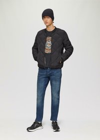 Zwarte gewatteerde bomberjack, grijs graphic t-shirt, blauwe jeans en zwarte sneakers. Model draagt een marineblauwe beanie. Eenvoudig, modern ontwerp.