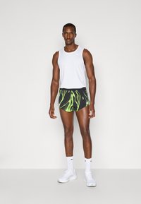 Balts bez piedurknēm topiņš un melni šorti ar zaļu abstraktu rakstu, apvienoti ar baltiem sporta apaviem un zeķēm. Vienkāršs dizains, sporta apģērbs.