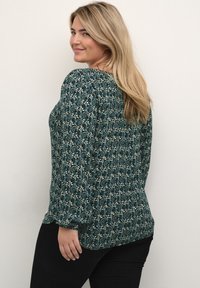 Grøn bluse med mønster og lange ærmer, der har blomsterdesigns og samlet talje. Bæres med sorte bukser.