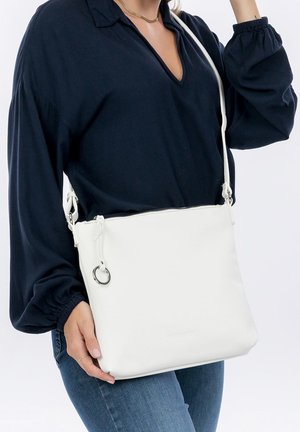 Kvinde iført mørk bluse med lange ærmer og jeans bærer en hvid læder crossbodytaske med en sølv ring detalje.