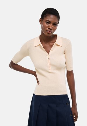 Femme portant un haut en maille beige à manches courtes avec col et une jupe plissée bleu marine ornée de boutons, debout avec une main sur la hanche.