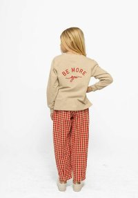 Béžová košeľa s dlhým rukávom s nápisom "BE MORE you" v červenej farbe na chrbte, v kombinácii s červeno-bielymi kockovanými nohavicami, obuté s béžovými topánkami.