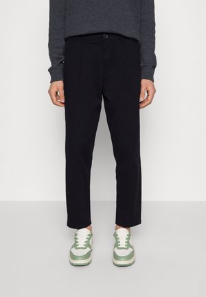 Personne portant un pantalon noir droit, un pull gris foncé et des baskets blanches avec des touches vertes, debout sur un sol clair devant un mur blanc.