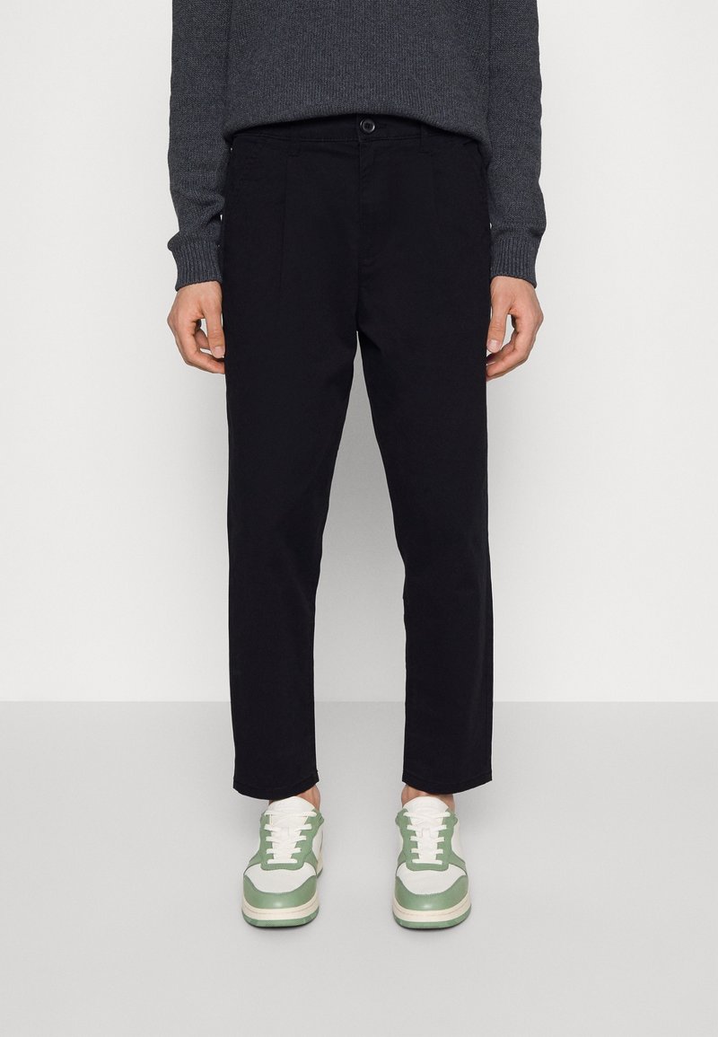 Personne portant un pantalon noir droit, un pull gris foncé et des baskets blanches avec des touches vertes, debout sur un sol clair devant un mur blanc.