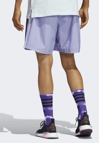 Pantaloni sportivi viola con fessure laterali, abbinati a calze tie-dye viola con strisce nere e sneakers nere con suole multicolori.