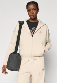 Bežinė zip-up megztinė su švelnia tekstūra, su subtiliu logotipu. Modelis laiko mažą juodą kryžminį krepšį su lygiu paviršiumi.