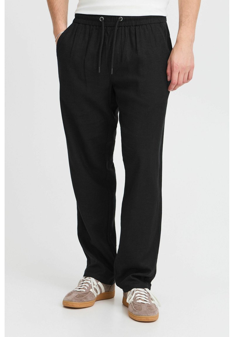 Black linen trousers with an elastic waistband, drawstring detail, and a relaxed fit. Paired with brown and white sneakers.