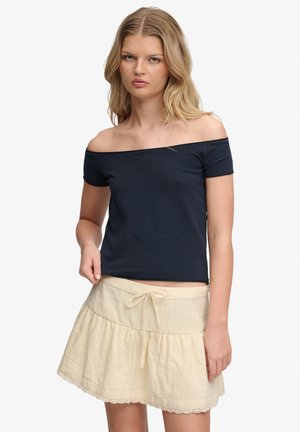 Blondynka w czarnym topie z odkrytymi ramionami i beżowej spódnicy z wiązaniem z przodu, stojąca na jednolitym białym tle.