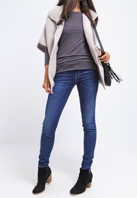 Cardigan en maille beige avec boutons, haut à manches longues gris, jeans slim bleu et bottines noires à talon épais.