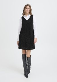 Crna haljina bez rukava s teksturiranim uzorkom, složena preko bijele košulje s gumbima. Model nosi crne kožne čizme do koljena.