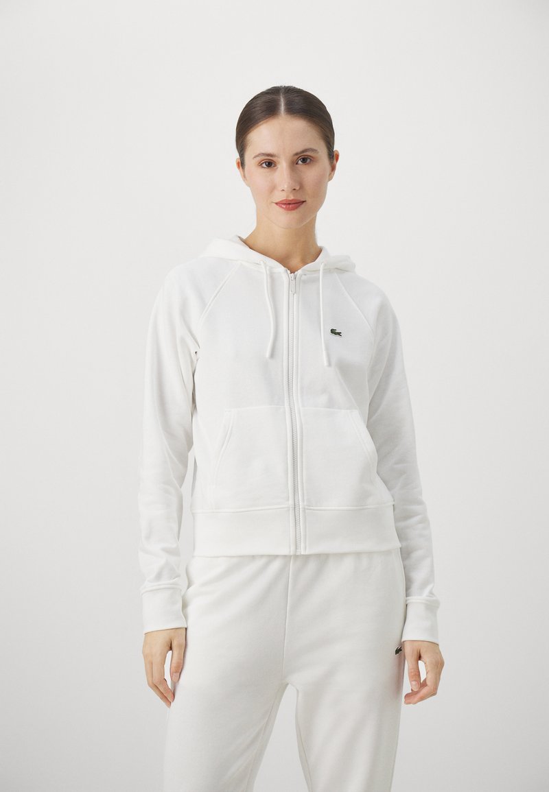 Bijela zip-up hoodie od mekog materijala s kapuljačom, rebrastim rukavima i kengur džepovima. Sadrži mali zeleni logo na lijevoj strani prsiju.